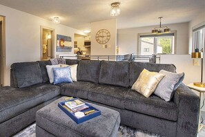 A modern living room with a grey sectional sofa, a coffee table with books, a large wall clock, and a kitchen in the background.