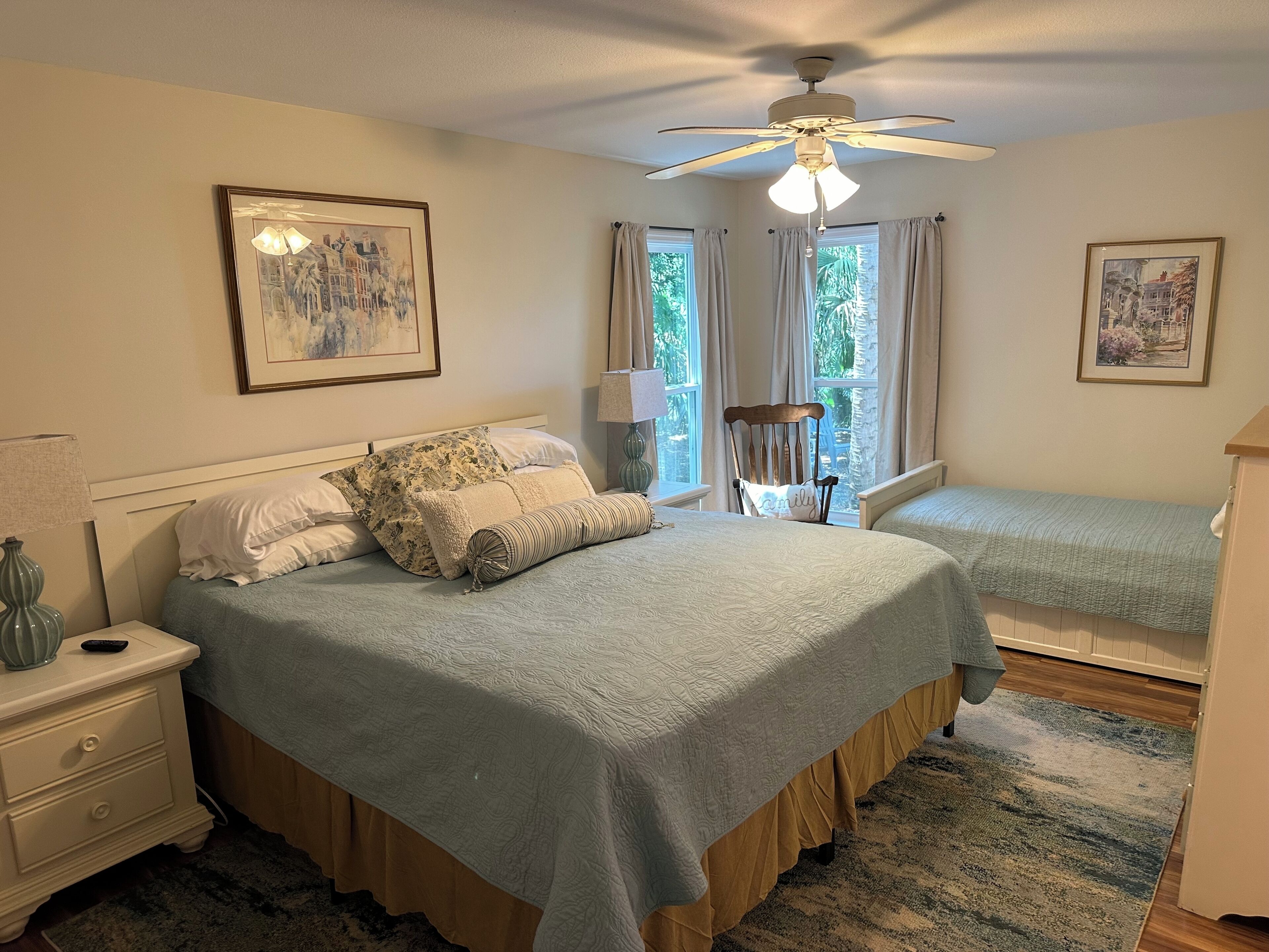 Une chambre à coucher comprenant un lit, deux tables de chevet, une chaise, un ventilateur de plafond et deux fenêtres avec des rideaux.