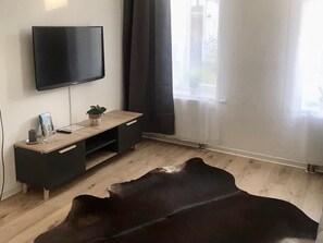A modern living room with a flat-screen TV mounted on the wall, a wooden coffee table, a black leather rug, and a window with sheer curtains. - Luxury Condo (Leipzig)