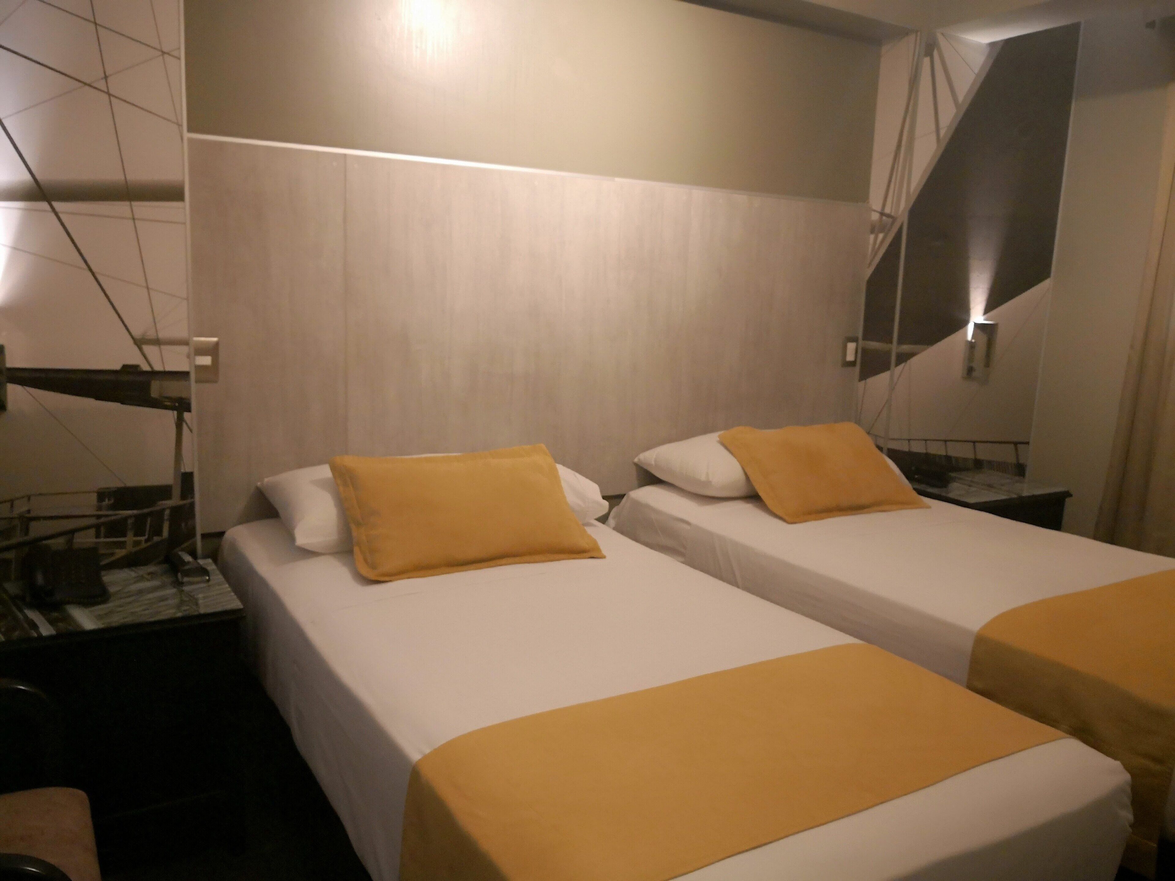 Habitación de hotel con dos camas, cada una con ropa de cama blanca y un cojín amarillo decorativo. La habitación presenta un diseño moderno con un espejo grande y un patrón geométrico en la pared.