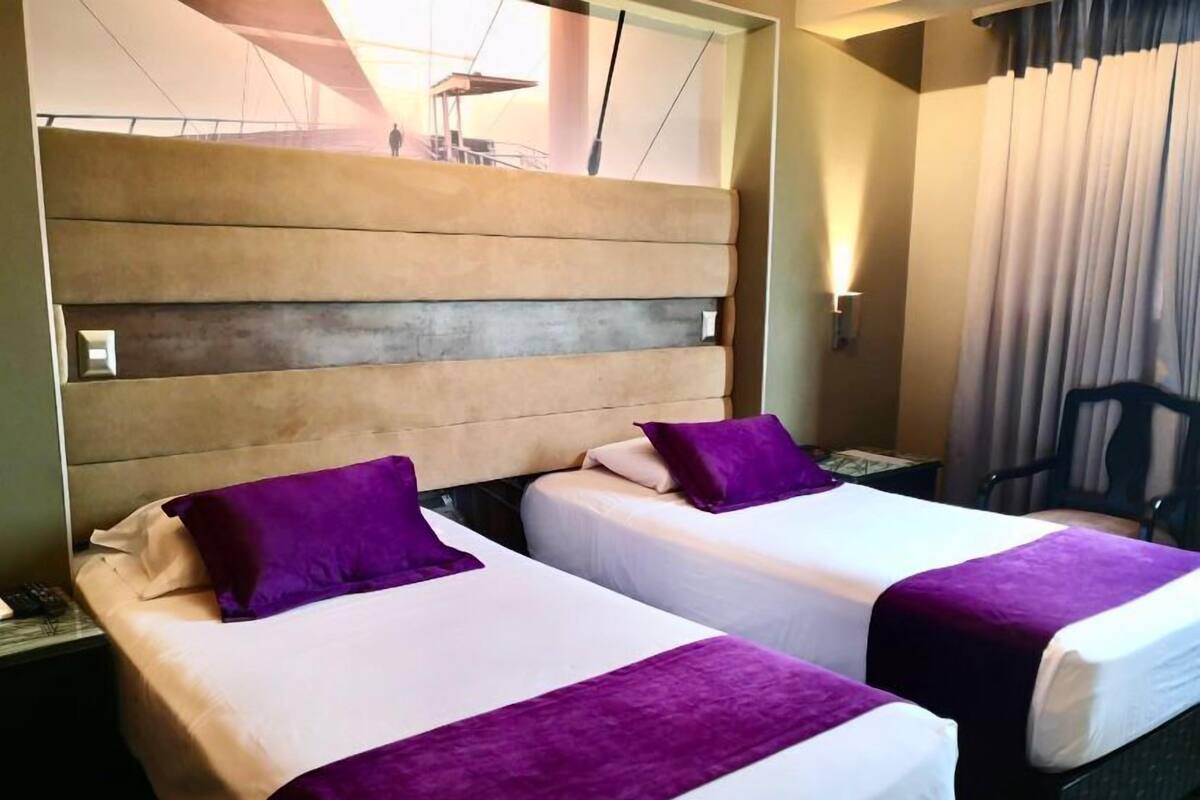 Habitación de hotel con dos camas, cada una con cabecera y almohada morada decorativa.