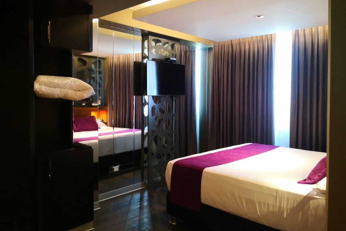 Habitación de hotel con cama, televisor, espejo y ventana con cortinas.