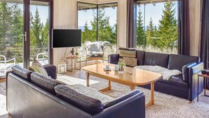 A modern living room with a large TV, comfortable seating, and a view of trees through large windows. - Beautiful home in øyer with sauna (Øyer)