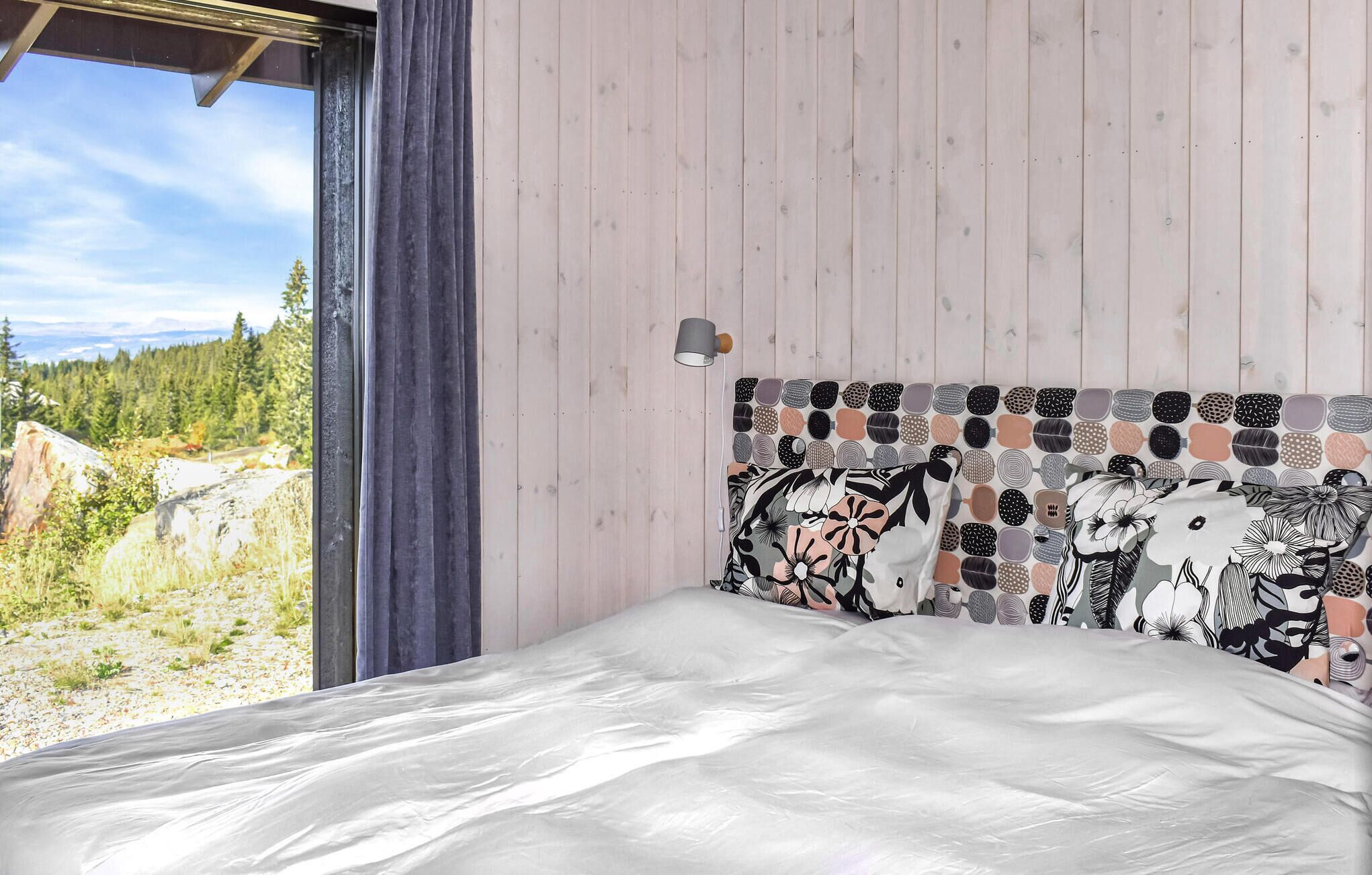 A bedroom with a bed, a headboard with a mosaic pattern, a window with a view of a forest, and a small wall-mounted lamp.
