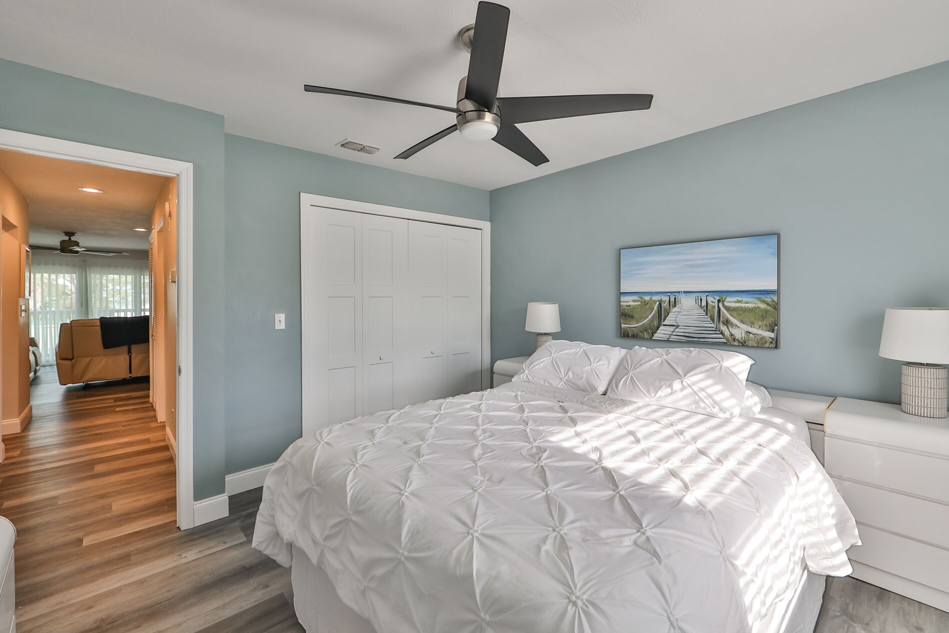 A bedroom with a bed, a ceiling fan, a white door, and a painting of a beach scene.