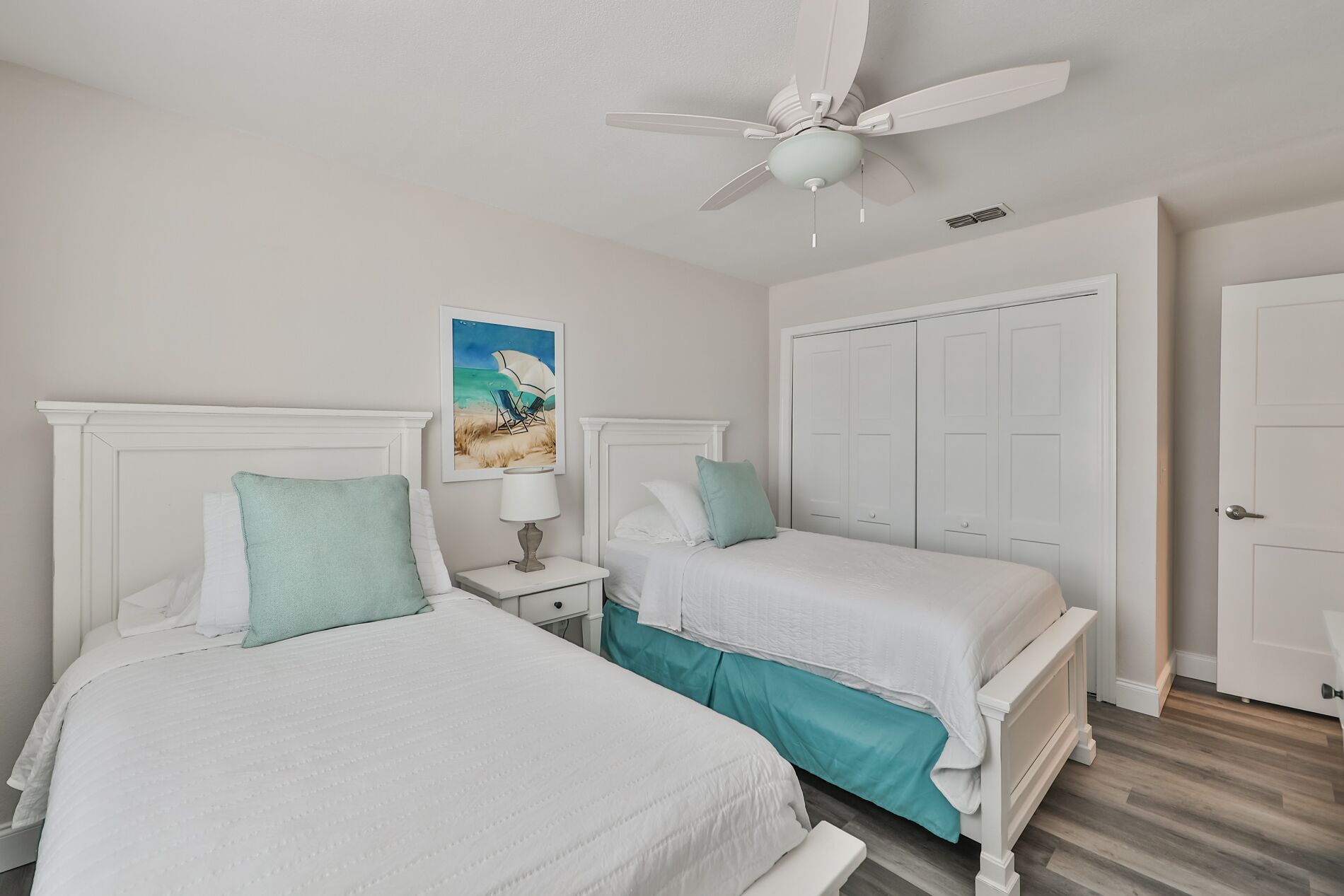 A bedroom with two single beds, a ceiling fan, a nightstand with a lamp, a painting of a beach scene, and a white wardrobe.
