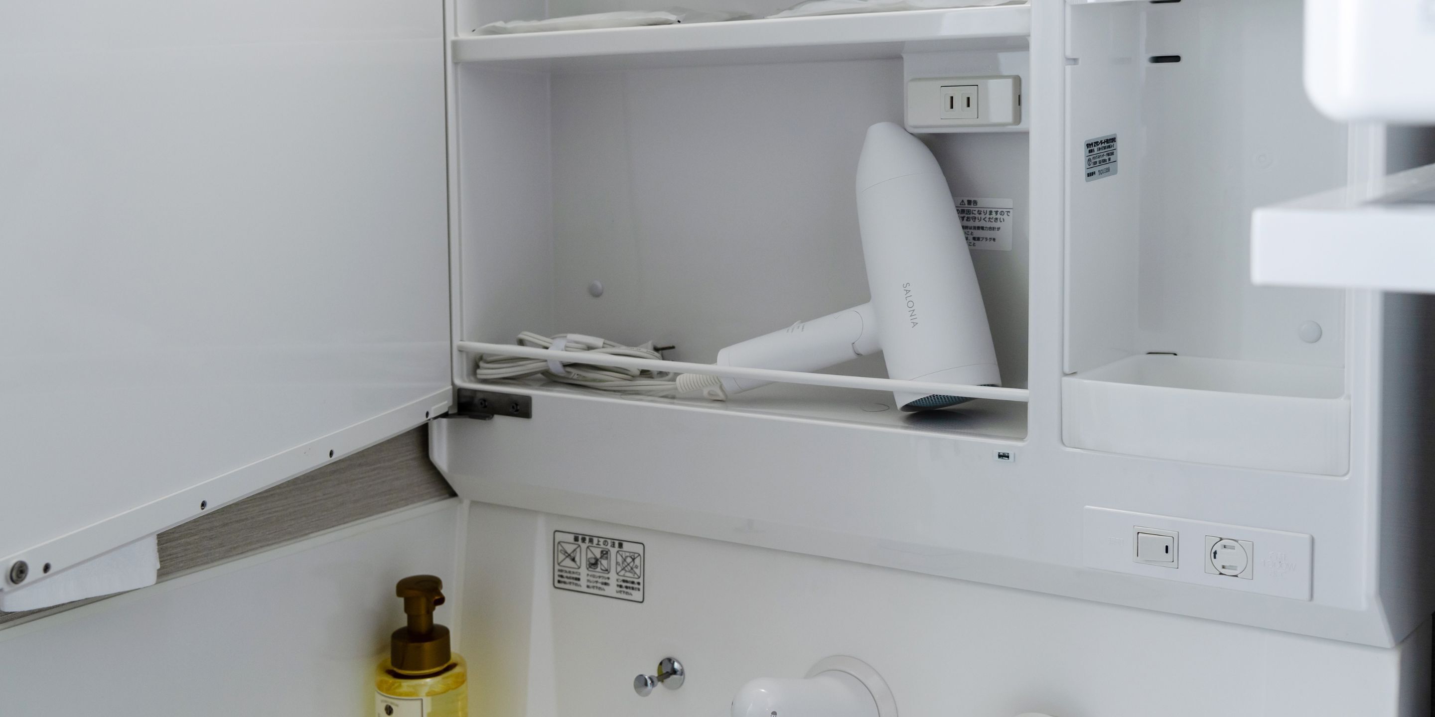 A white bathroom with a hairdryer on a shelf, a bottle of hand soap, and a toilet paper holder.