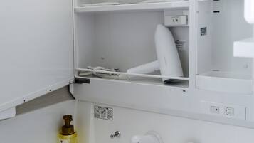 A white bathroom with a hairdryer on a shelf, a bottle of hand soap, and a toilet paper holder.