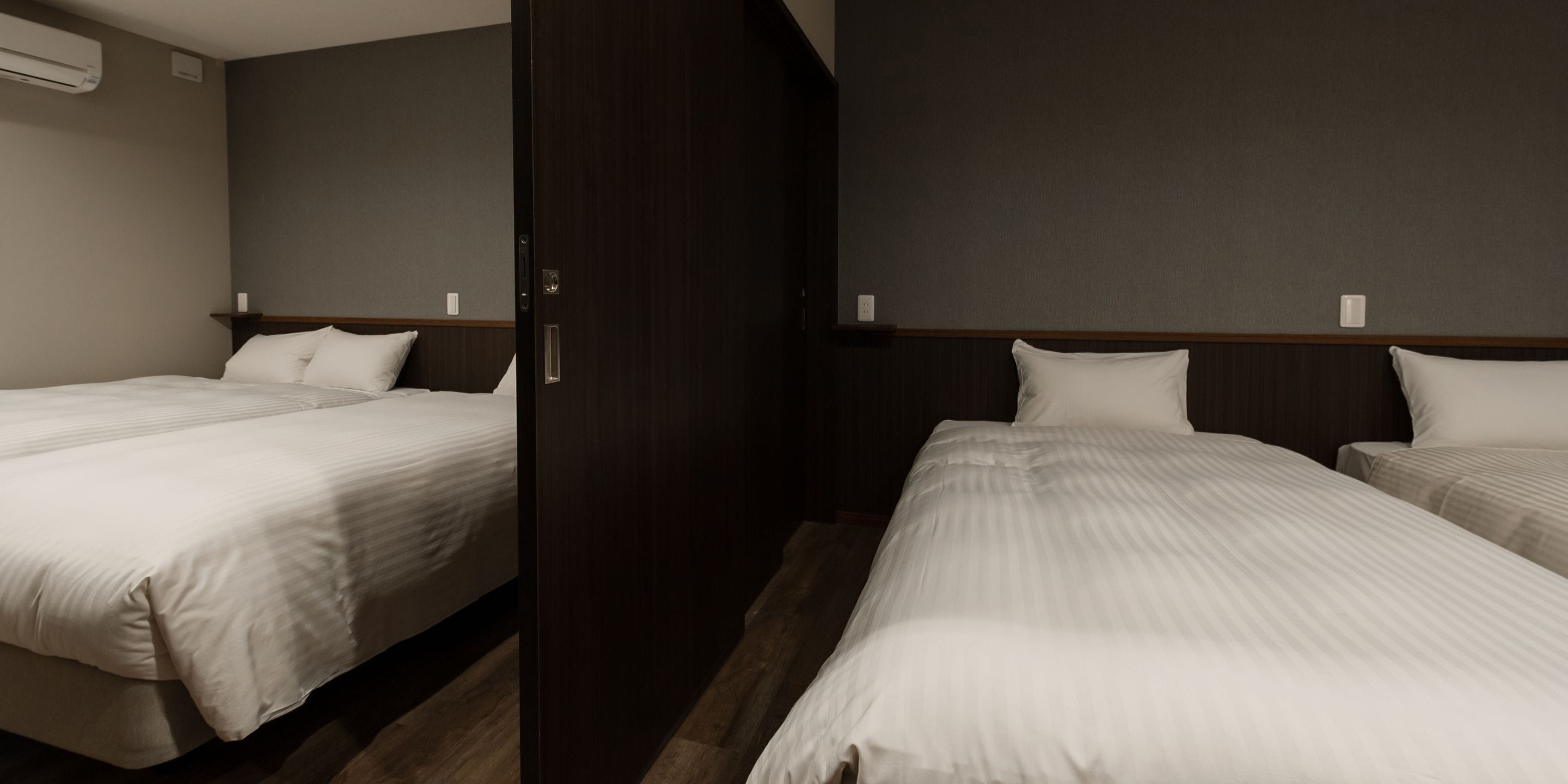 A hotel room with two beds, a wooden wardrobe, and a neutral-colored wall.