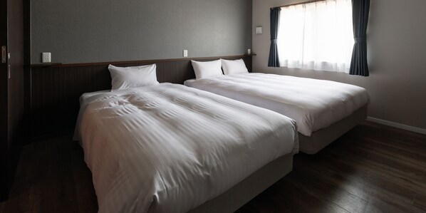A hotel room with two single beds, a wooden headboard, a grey wall, and a window with curtains.