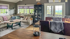 A cozy living room with a sofa, a wooden coffee table, and a dining table. There is a glass cabinet with dishes and a view of the outdoors through large windows. - Amazing home in Rena with sauna (Rena)
