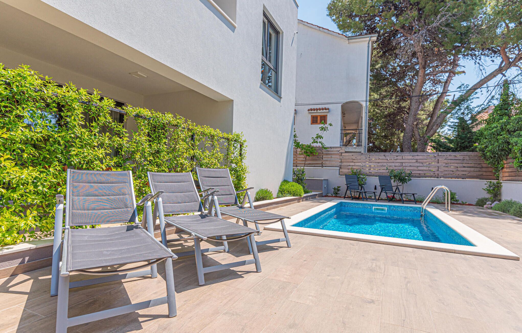 Zona junto a la piscina con sillas de descanso, piscina y un edificio blanco con terraza de madera y vegetación.