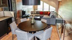 Ein modernes Wohnzimmer mit einem runden Esstisch, grau gepolsterten Stühlen, einer schwarzen Stehlampe, einer Couch mit bunten Kissen und einem Glasgeländer mit Blick auf eine Holzterrasse.