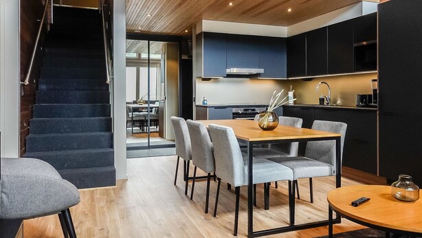 Eine moderne Küche mit dunklen Schränken und einer Holzbalkendecke, ein Essbereich mit grauen Stühlen und einem Holztisch sowie eine Treppe, die zu einer weiteren Ebene führt.