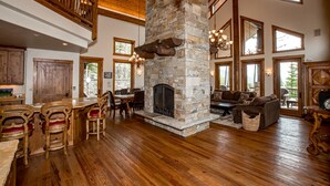 A spacious living area with a stone fireplace, wooden flooring, and a high ceiling with exposed beams.