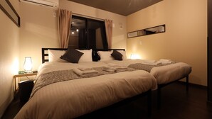 A hotel room with two beds, a nightstand with a lamp, an air conditioner, a window with curtains, and a mirror on the wall. - SORAPIA Villa Mt.Fuji Front (Fujikawaguchiko)