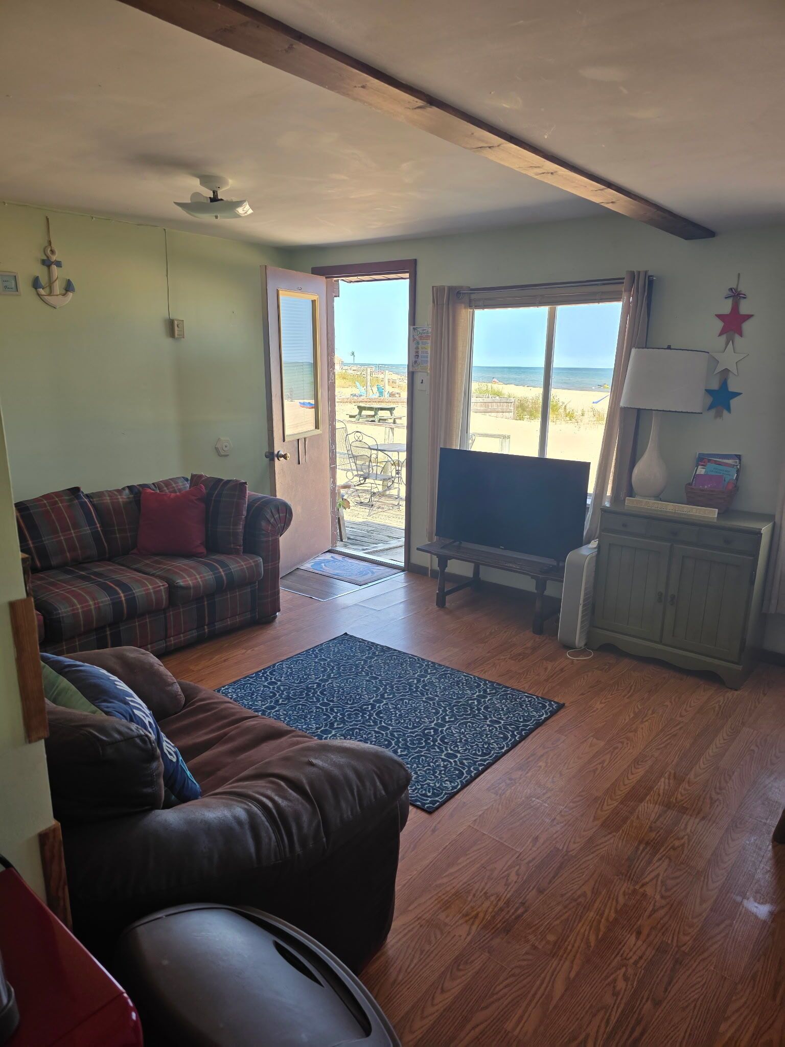 A living room with a sofa, a television, a wooden floor, and a view of the beach through a door.