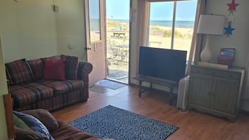 A living room with a sofa, a television, a wooden floor, and a view of the beach through a door.