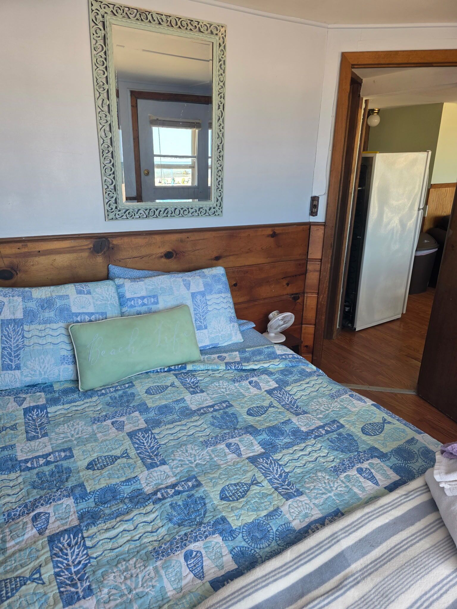 Un lit avec une couvre-lit à motifs bleus et blancs, un oreiller orné du texte « Beach Life », et une tête de lit en bois.