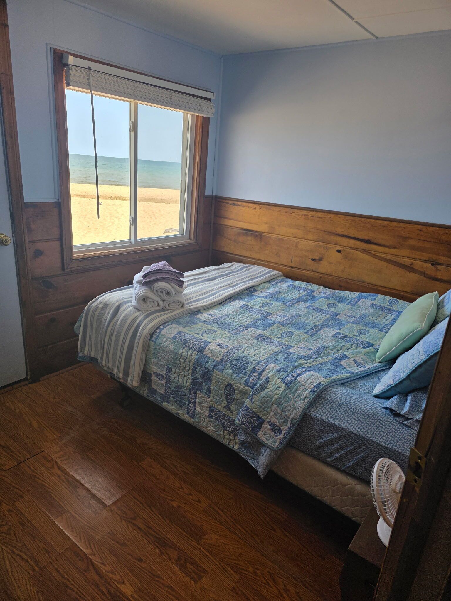 A bedroom with a wooden headboard, a bed with a patterned quilt, a window with a view of a beach, and a wooden floor.