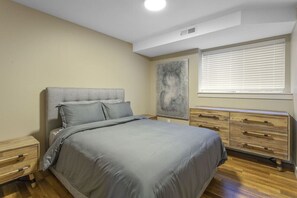 A bedroom with a bed, a nightstand, a dresser, a window with blinds, and a wall-mounted artwork.