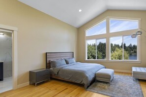 A bedroom with a large bed, a nightstand, a bench, and a view of a forest through large windows.