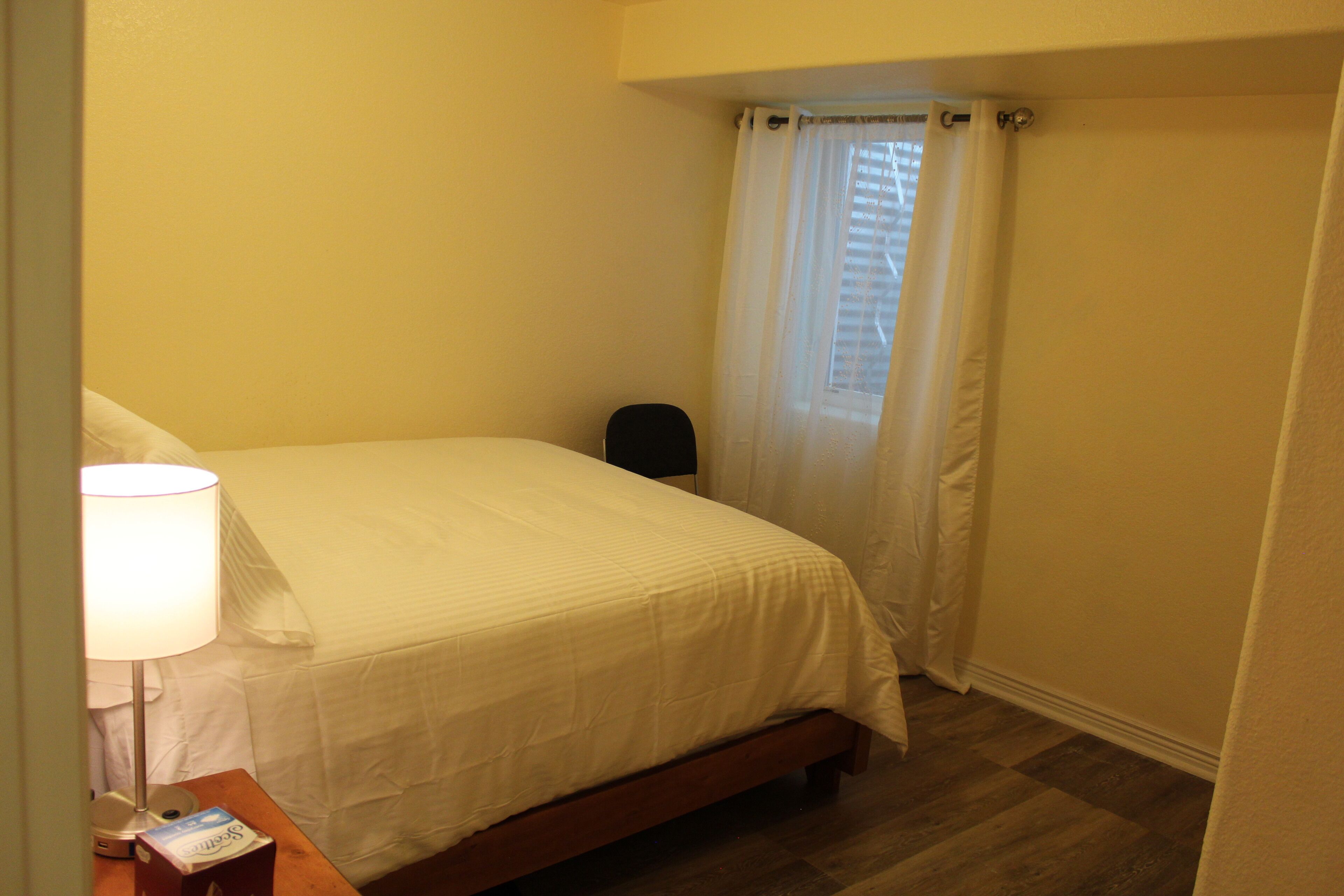 Una camera da letto piccola e curata, con un letto singolo, un comodino con una lampada e una finestra con tende.