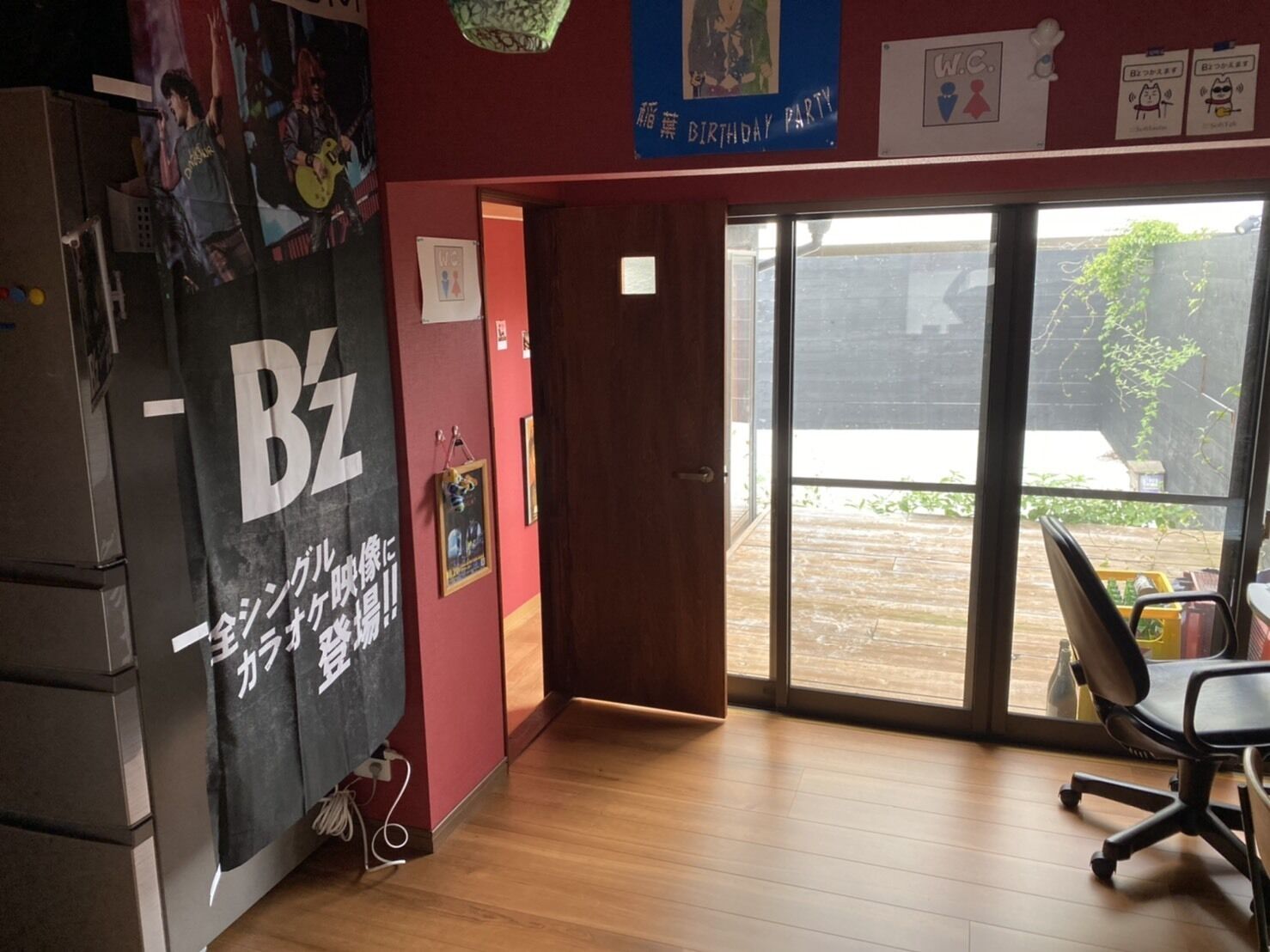 Una habitación con piso de madera, un mueble negro con el logotipo 'Bz', una puerta que da al exterior y una ventana con vista a una terraza de madera.
