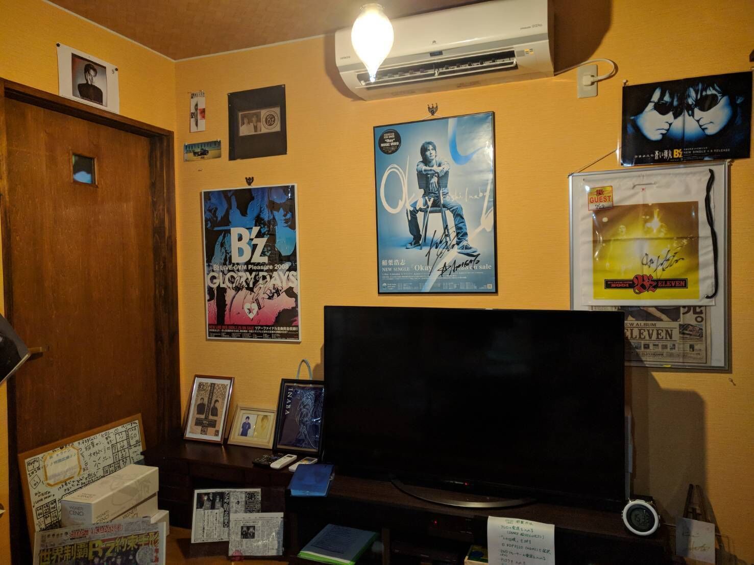 Une pièce dotée d’un grand téléviseur à écran plat, de posters accrochés au mur, d’une porte en bois et d’un petit bureau sur lequel se trouvent divers objets.