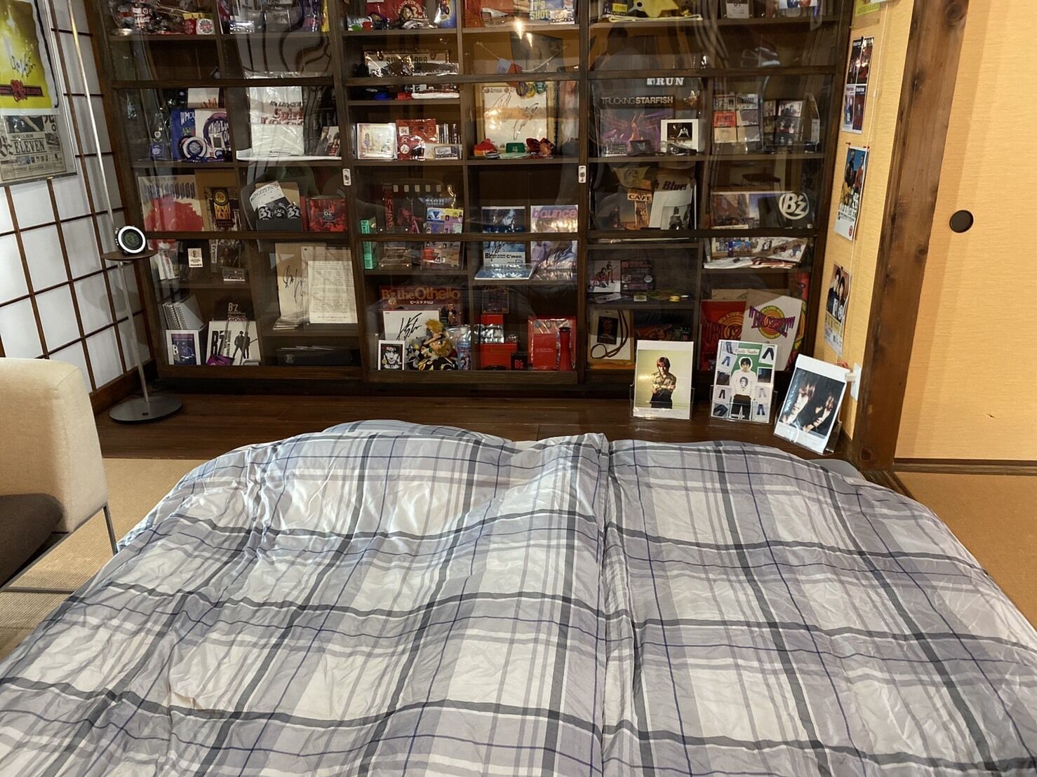 A bedroom with a plaid bedspread, a bookshelf filled with various items, and a small desk with a lamp.