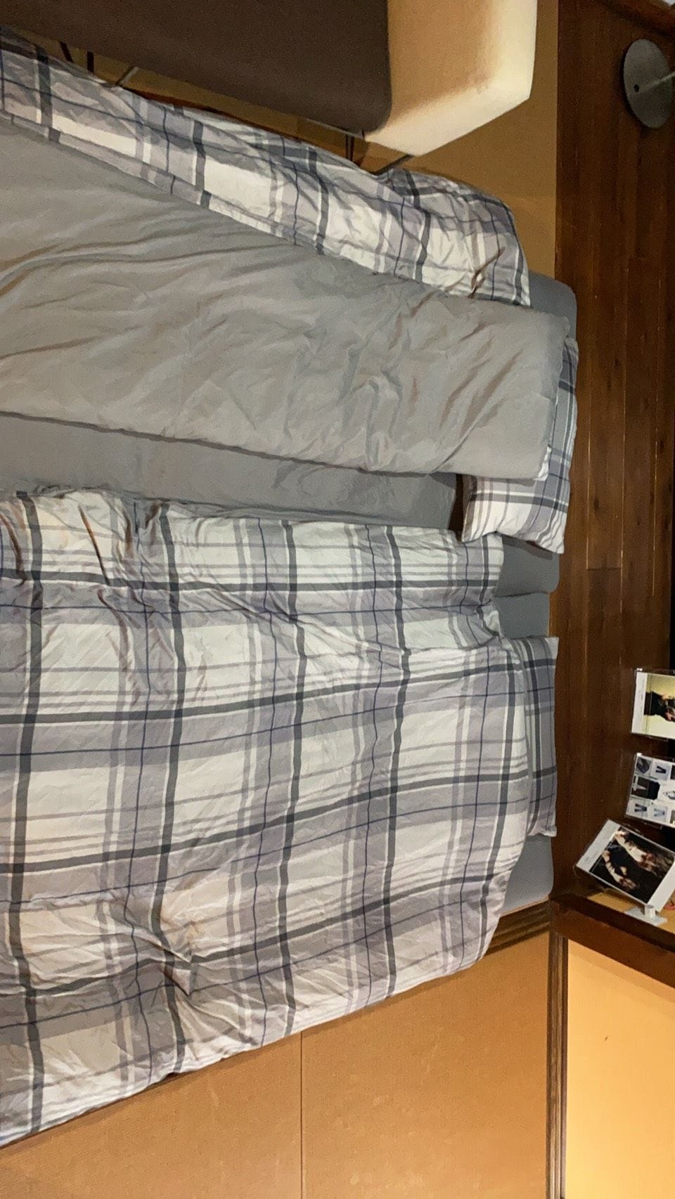 Una cama con una manta y almohadas a cuadros, un cabecero de madera y una mesita con una revista.