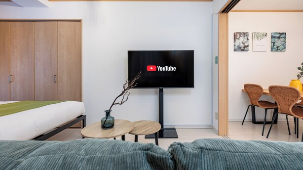 A modern hotel room with a bed, a TV mounted on the wall, a small table with chairs, and a dining area with wicker chairs and a yellow vase.