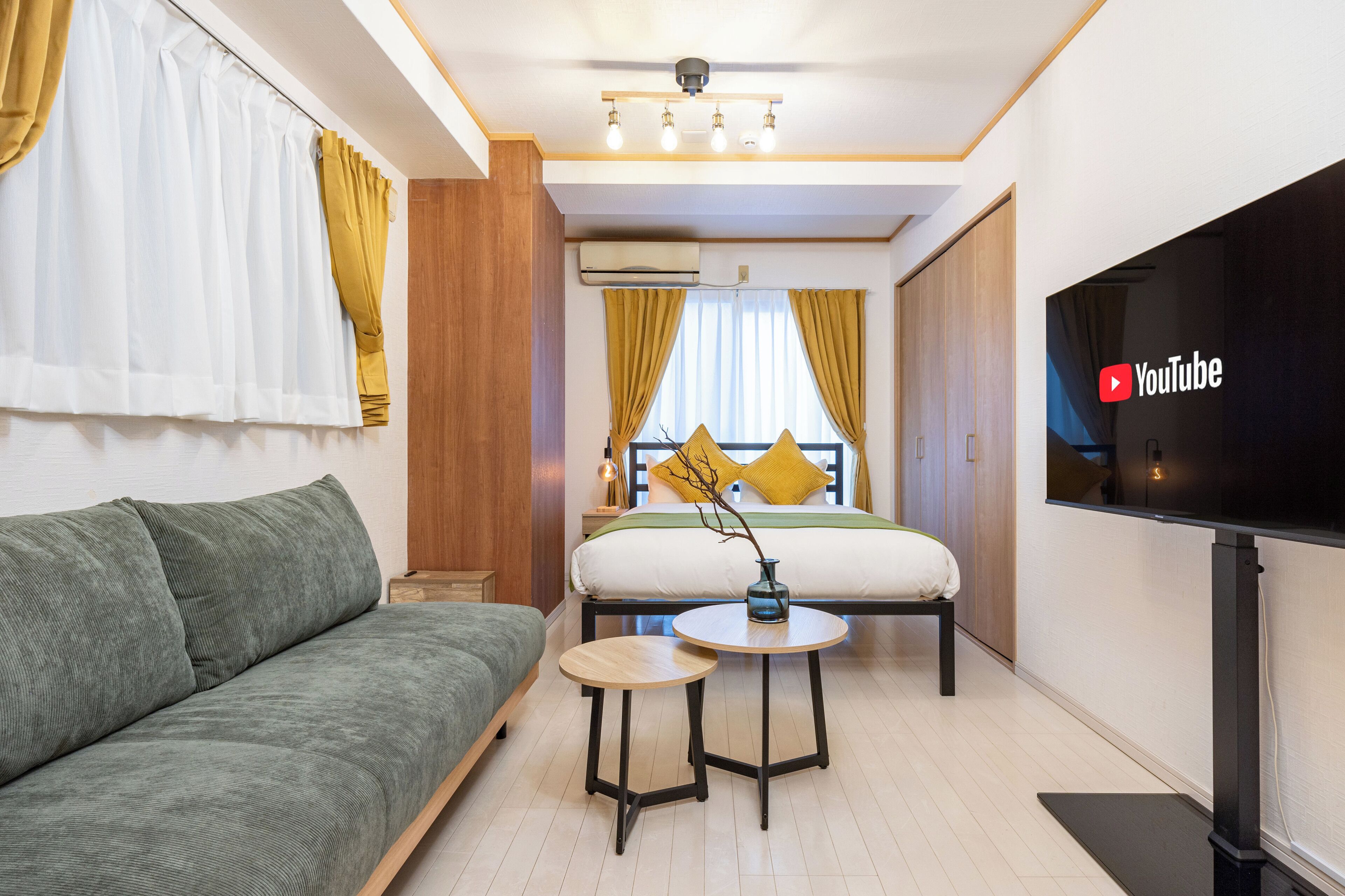 A modern hotel room with a green sofa, a small round table, a bed with yellow pillows, and a TV on the wall.