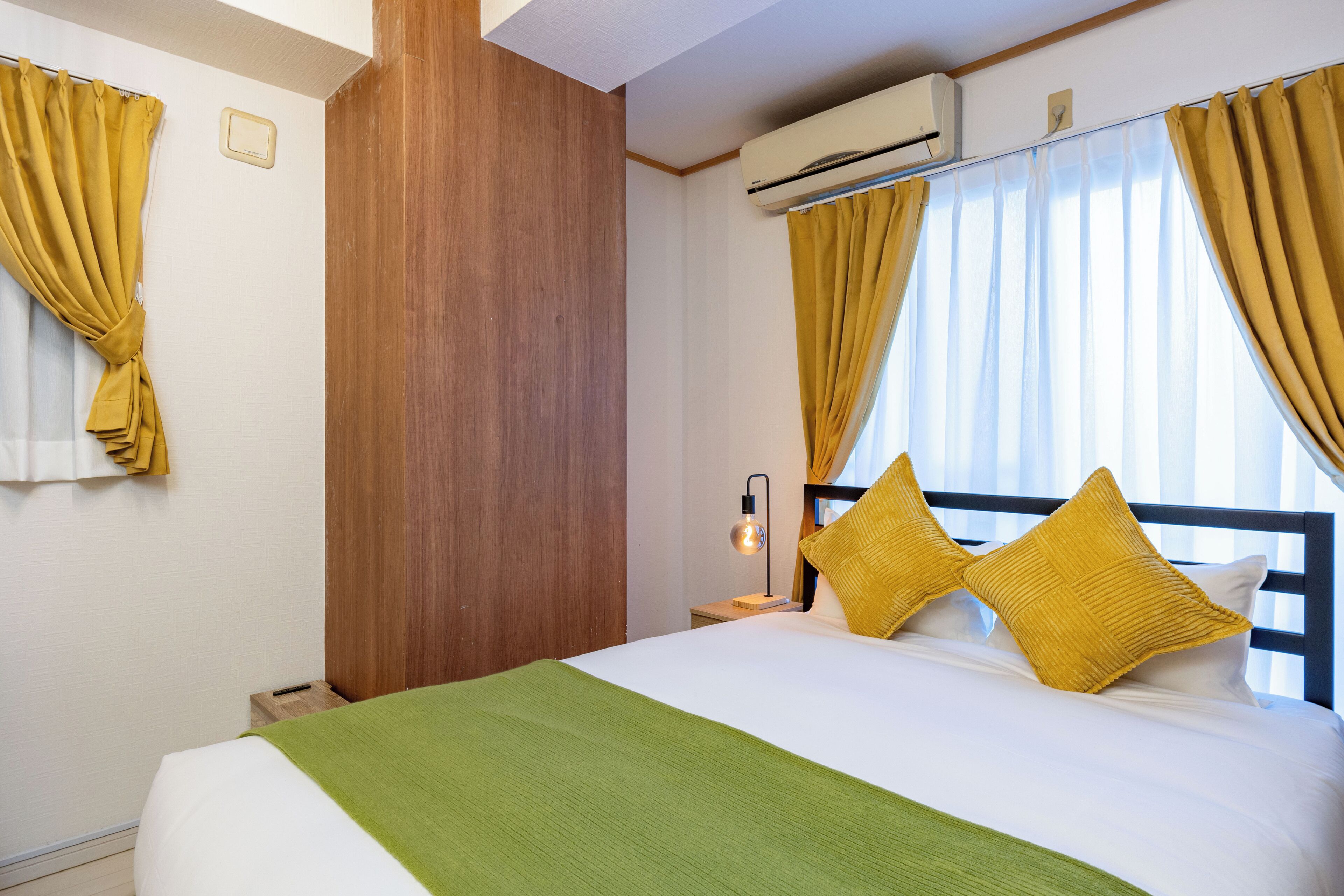A hotel room with a bed, yellow curtains, an air conditioning unit, and a nightstand with a lamp.