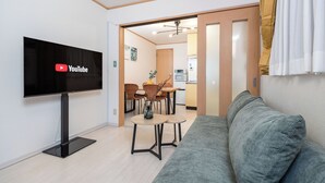 A modern living room with a flat-screen TV, a grey sofa, and a dining area with wicker chairs and a small table.