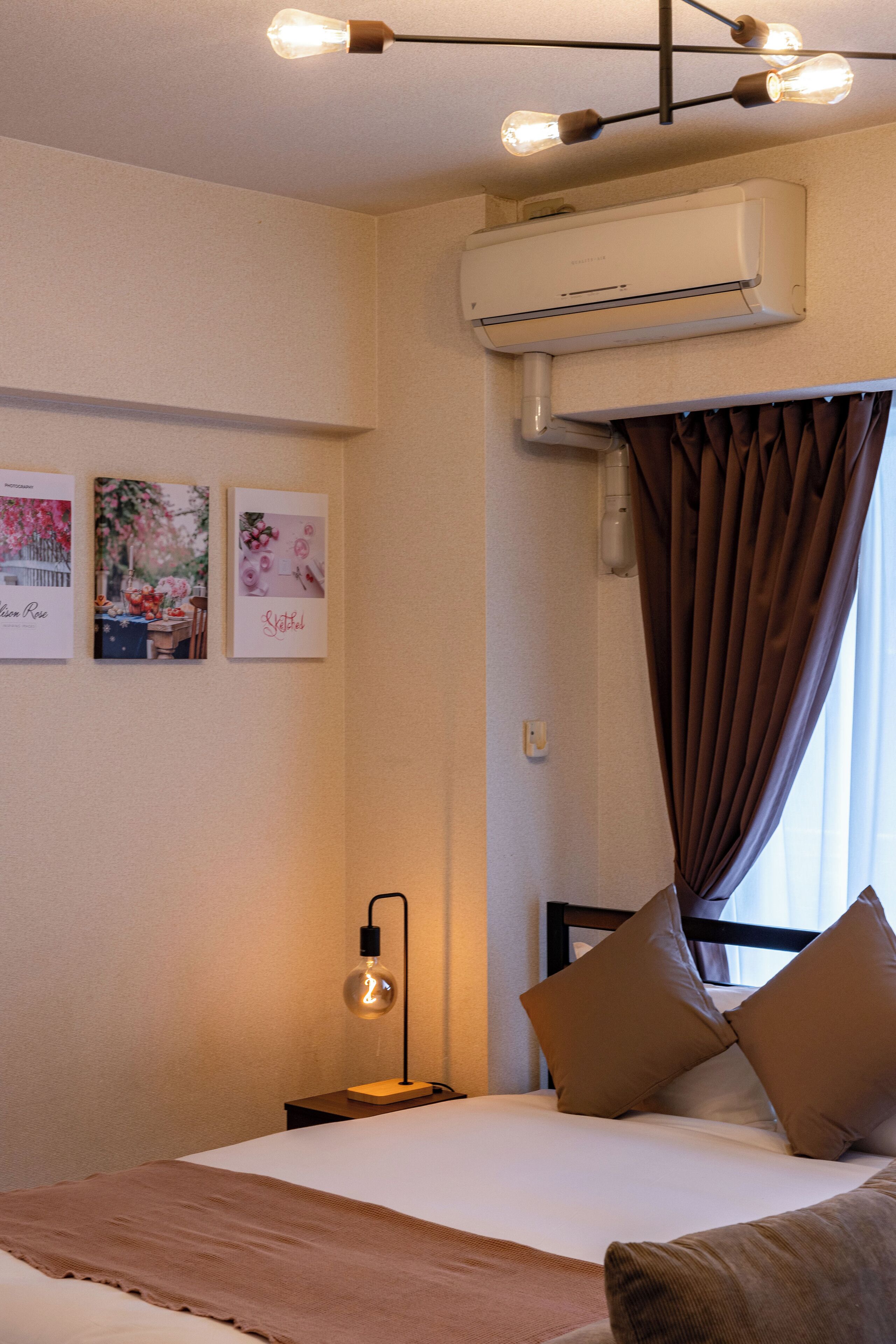 A bedroom with a bed, a bedside table with a lamp, a wall-mounted air conditioner, and framed pictures on the wall.