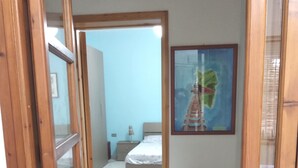 A room with a bed, a chair, a small table, a painting, and a wicker basket. - Base Camp Ogliastra - Flat Green  (Santa Maria Navarrese)