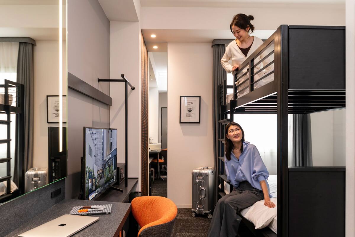 Quarto de hotel moderno com cama beliche, uma escrivaninha com um laptop, televisão e mala.