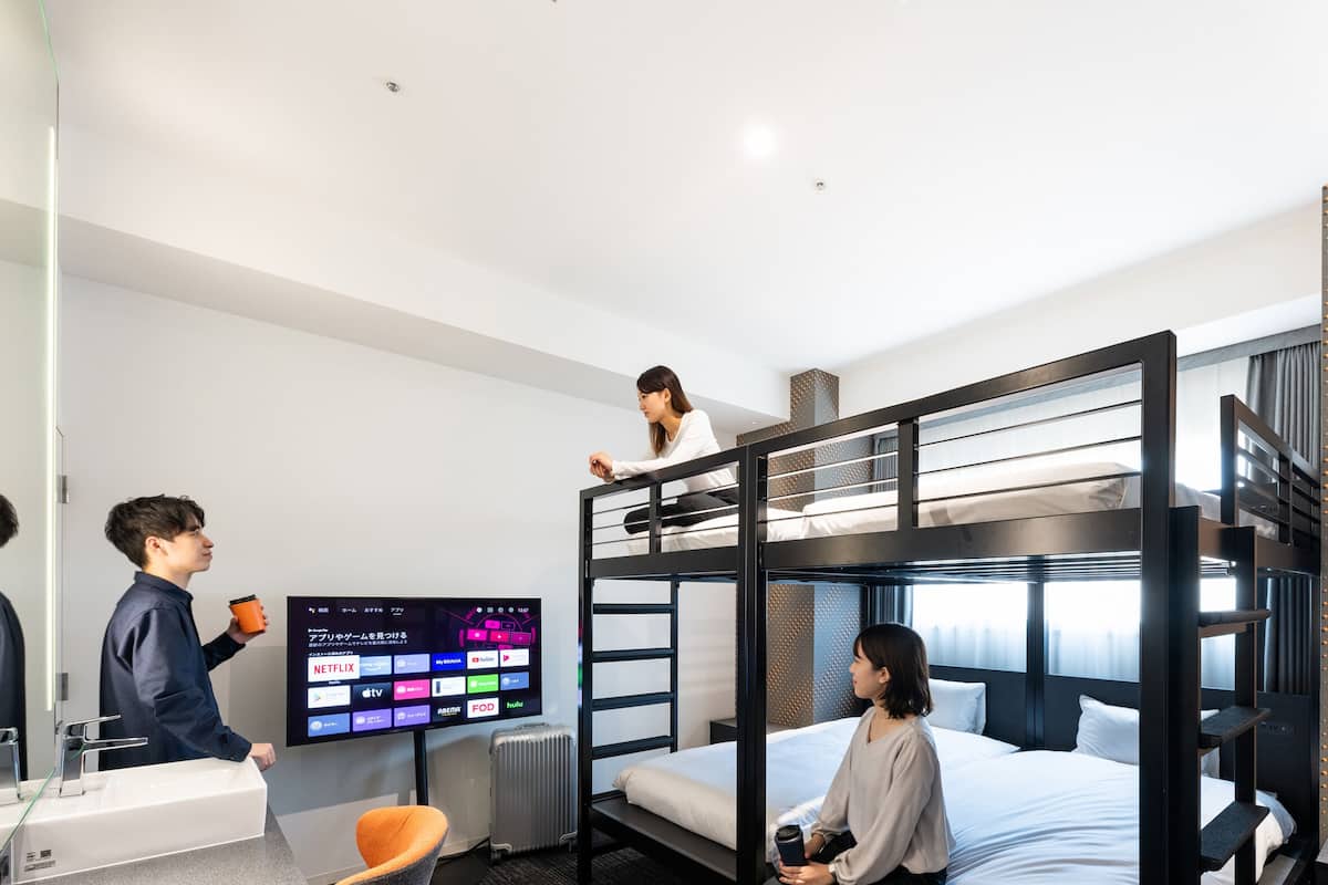 Quarto com beliche, televisão exibindo diversos serviços de streaming, escrivaninha com cadeira e mala.
