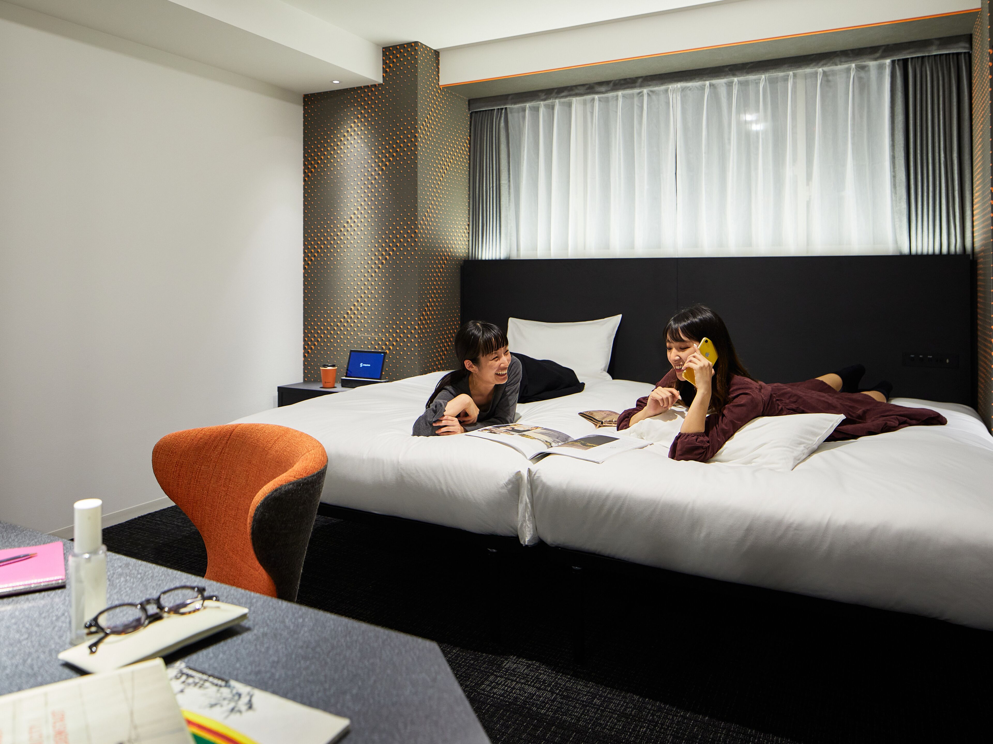 A hotel room with a bed, a nightstand with a laptop, a chair, and a table with a notebook and glasses.