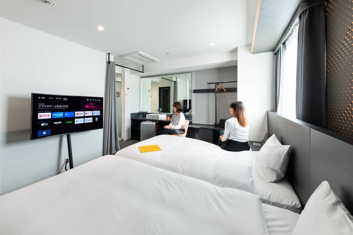 Quarto de hotel moderno com duas camas, televisão de tela plana exibindo serviços de streaming, espelho e janela com cortinas.