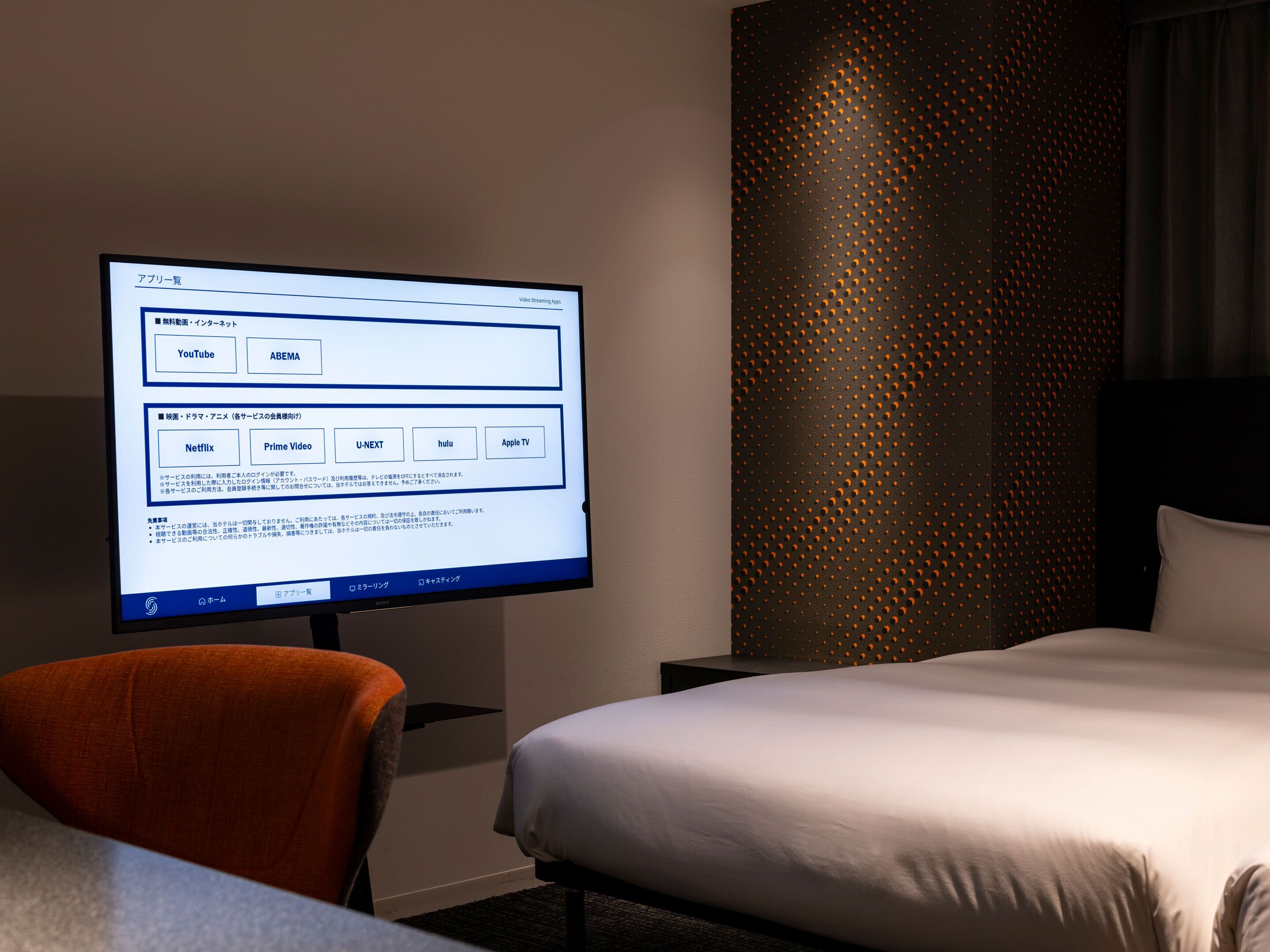A hotel room with a bed, a TV displaying a streaming service interface, and a wall with a perforated pattern.