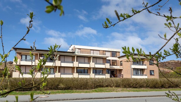 A modern multi-story building with balconies and large windows, surrounded by greenery and a clear sky. - Lister Dünennest, Deutschland (List)