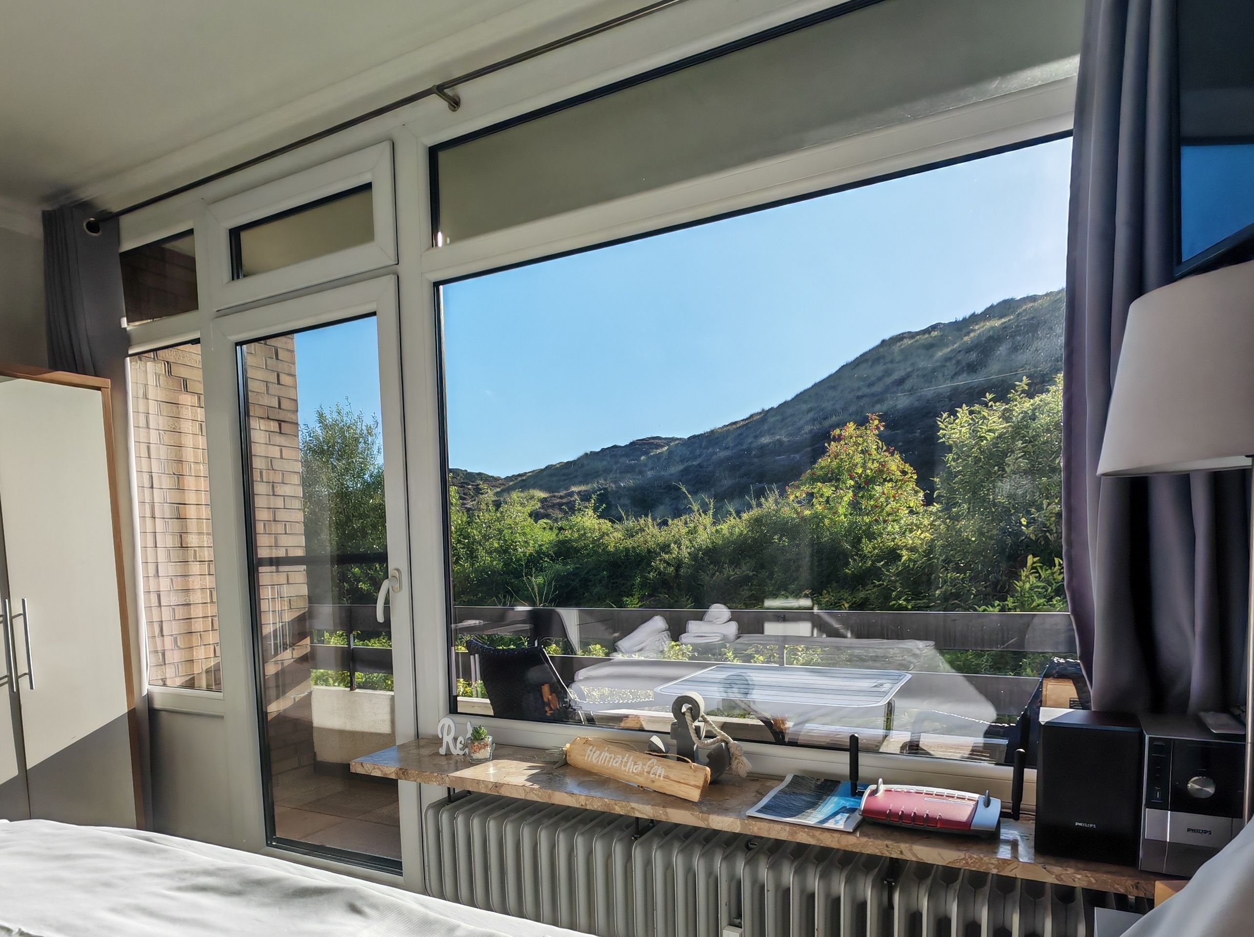 Ein Hotelzimmer mit großem Fenster, einem Bett, einem Schreibtisch mit Computer und Blick auf eine gebirgige Landschaft.