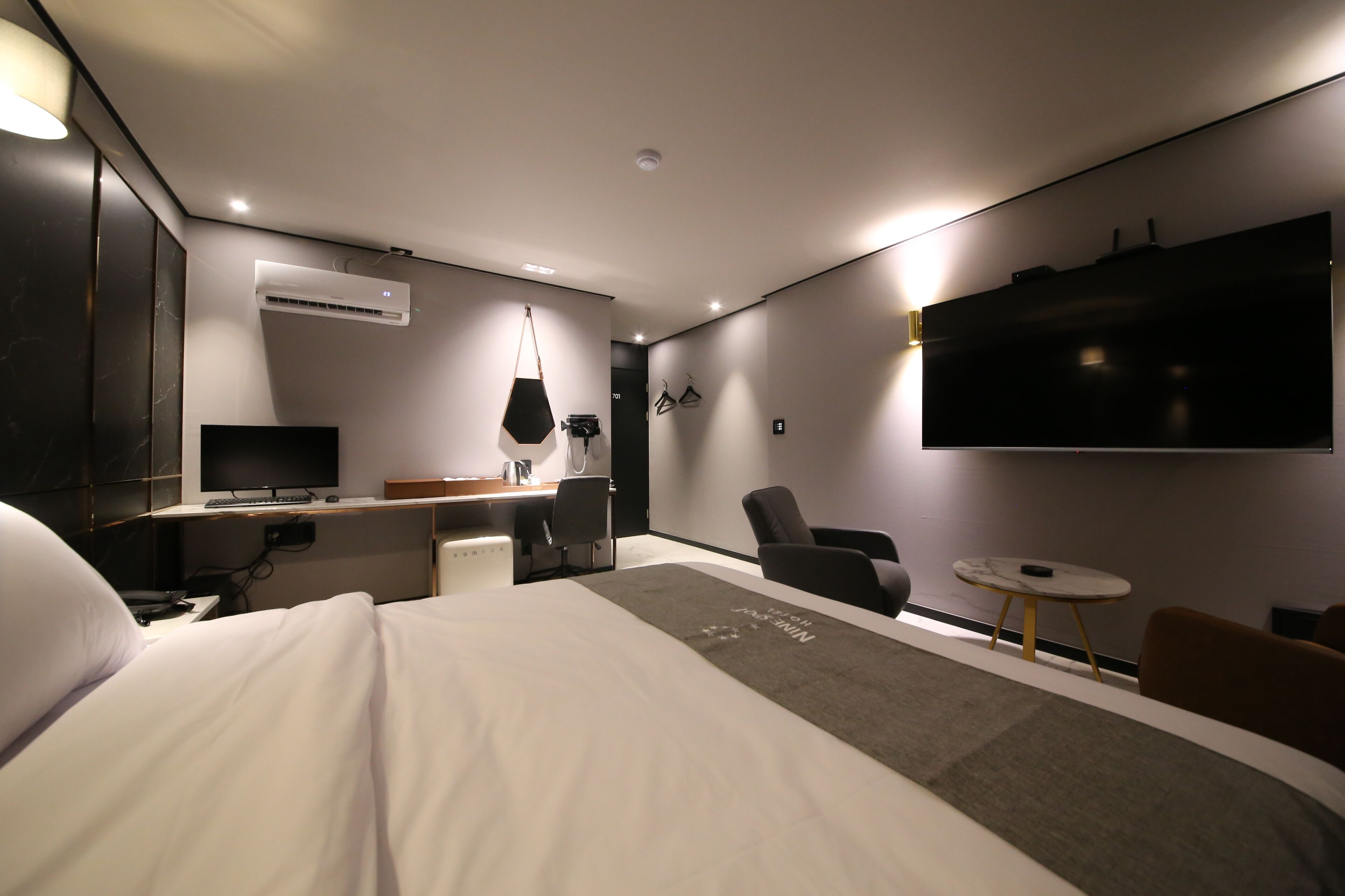 Et moderne hotelværelse med en stor seng, et skrivebord med computer, et fjernsyn og et siddeområde med stole og et lille bord.