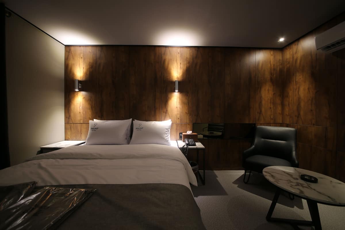 Ein modernes Hotelzimmer mit einer Wand in Holzoptik, einem Bett mit weißer Bettwäsche, einem schwarzen Ledersessel und einem Marmortisch.