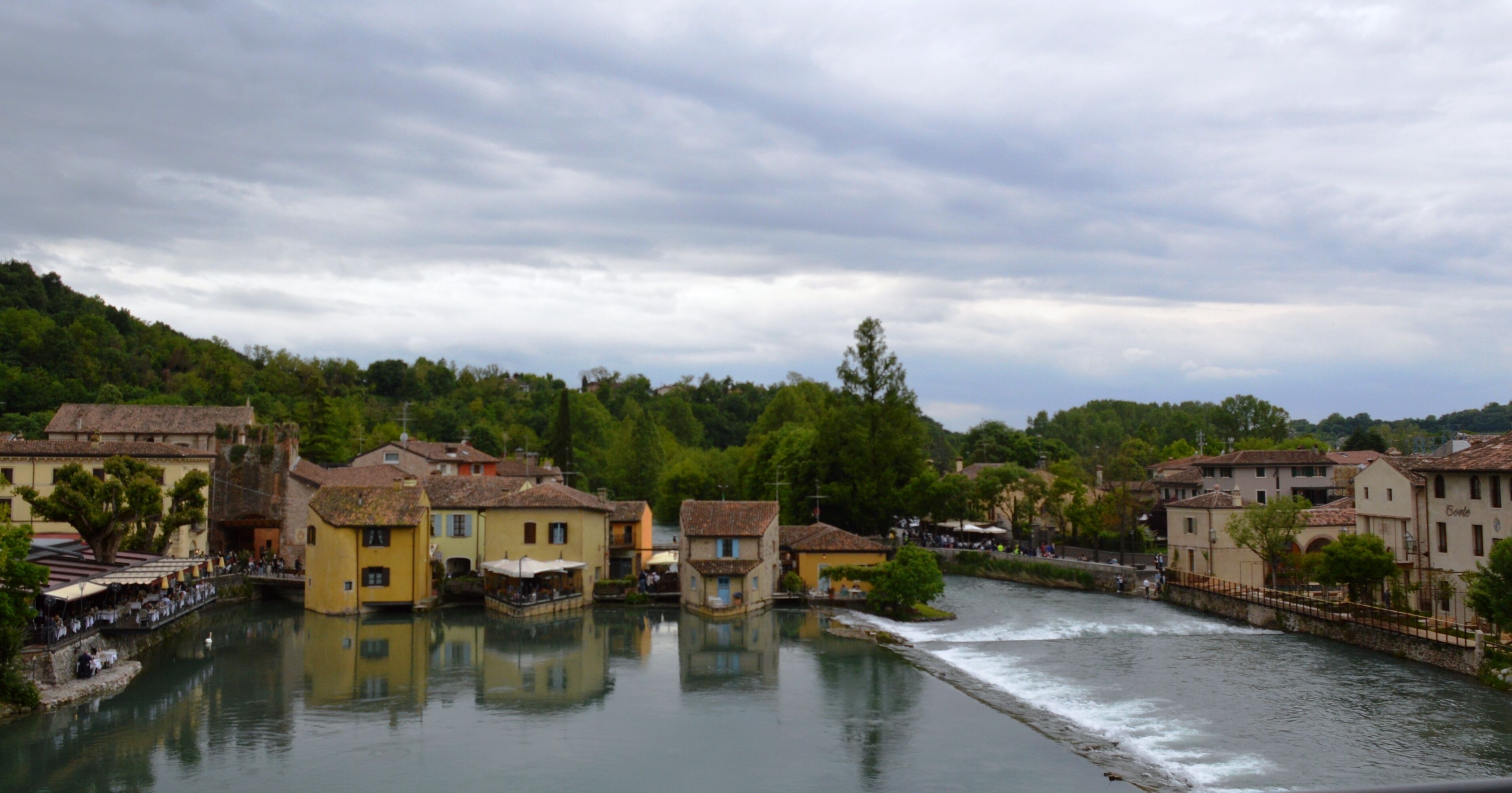 Une ville riveraine, avec des bâtiments qui bordent le cours d’eau, un pont et une structure qui ressemble à un château.