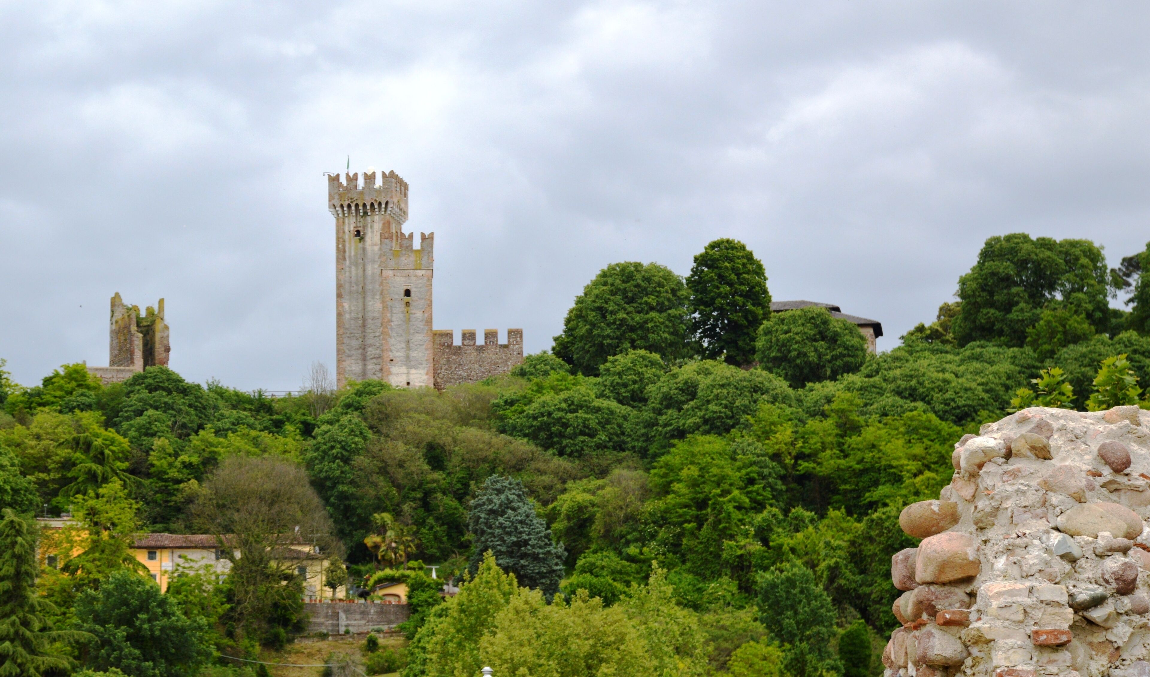 A historic stone tower with battlements, surrounded by lush greenery and a small building with a tiled roof.