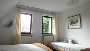 A room with two single beds, a wooden bench, a ceiling light, a framed picture on the wall, and two windows with curtains.