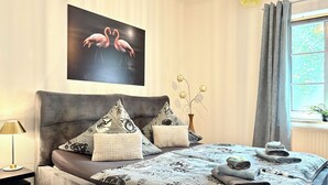 Makuuhuone, jossa on sänky, tyynyjä, yöpöytä, lamppu, maljakko kukkineen ja flamingoja esittävä taulu.